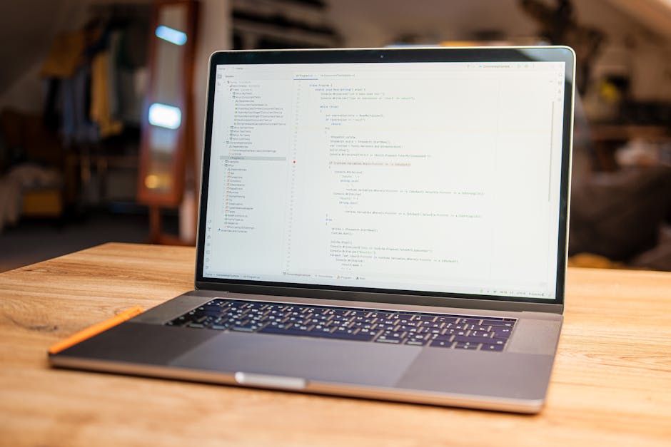 A silver MacBook laptop displaying lines of source code on its screen, placed on a wooden desk with a natural grain texture. The background is softly blurred, revealing a cozy indoor setting with warm